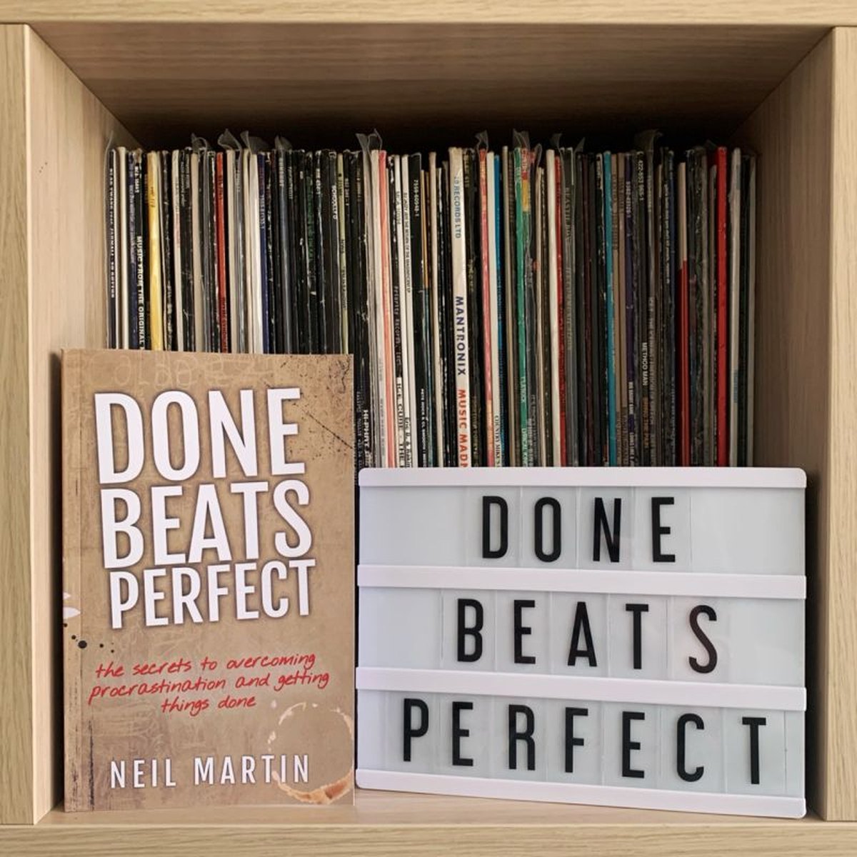 A copy of Neil Martin's book Done Beats Perfect next to a light box sign reading Done Beats Perfect, in front of a shelf of vinyl records