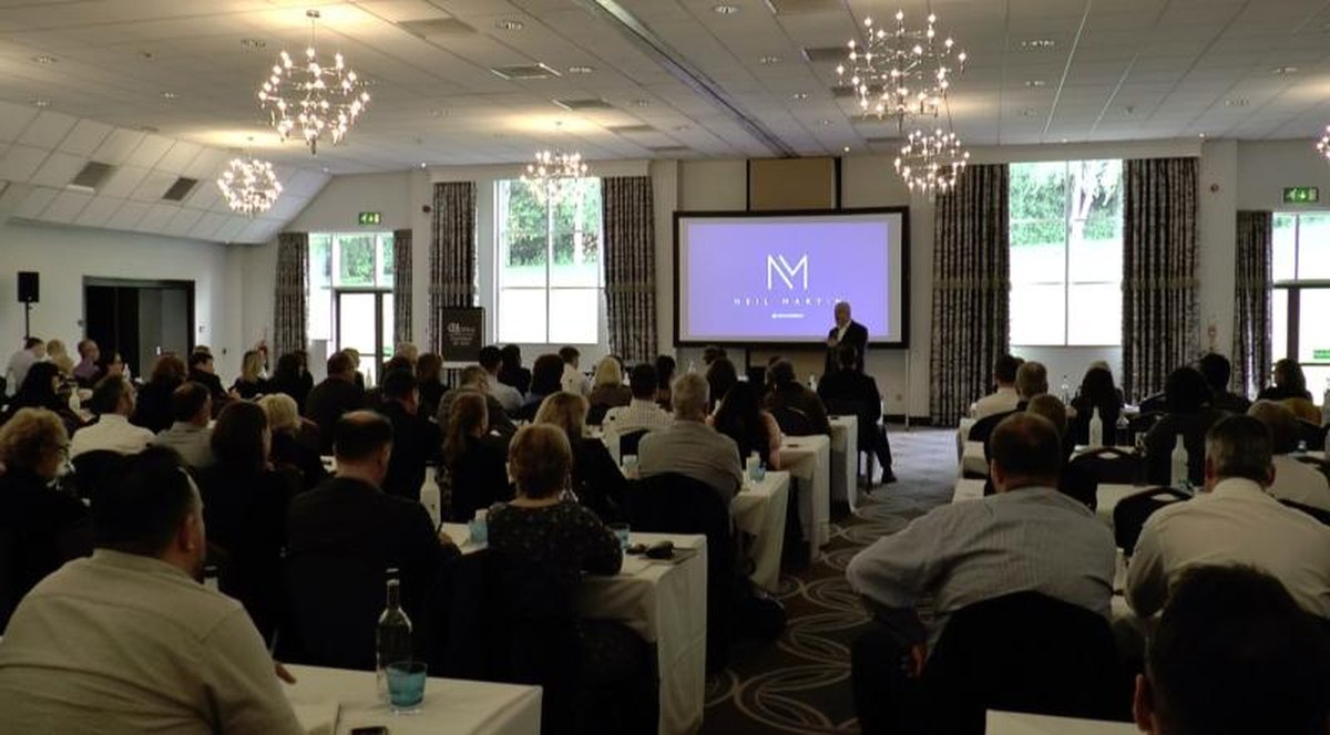 A packed conference room with chandeliers and an audience at round tables, facing a speaker beside a screen showing the Neil Martin logo