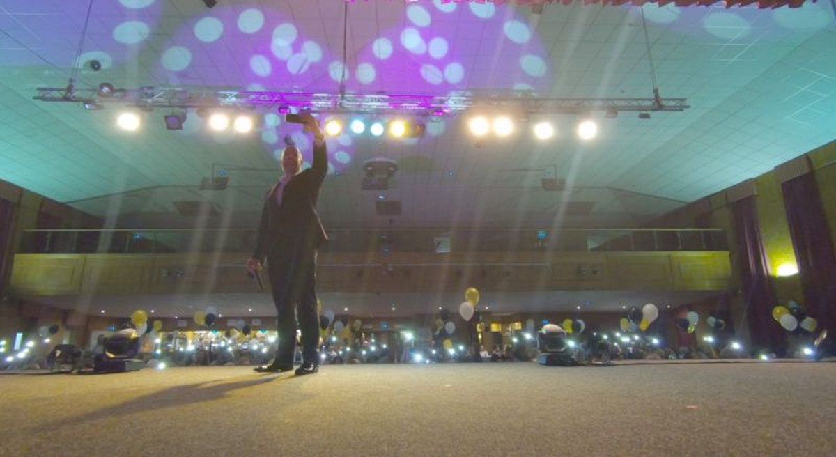 Neil Martin on a big stage under coloured lights, holding his phone up for a selfie with a room full of tables and balloons behind him